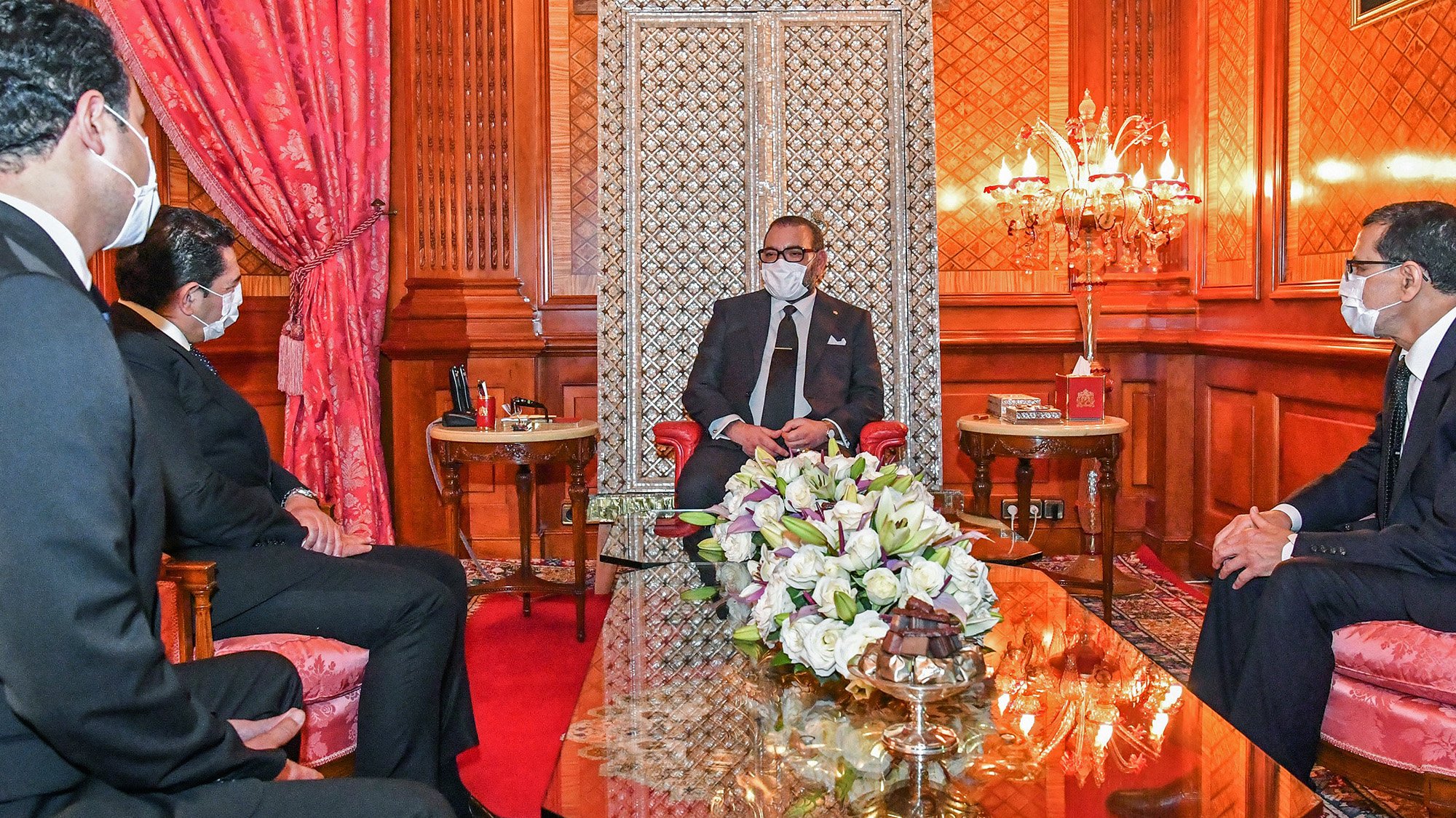 در این تصویر، یک جلسه رسمی در یک اتاق مجلل نمایش داده شده است. در مرکز تصویر، یک مرد (که به نظر میرسد مقام بلندپایهای است) بر روی صندلی نشسته و با لباسی رسمی و ماسک به چهره دارد. سه مرد دیگر در سمت چپ و راست او نشستهاند و به او گوش میدهند. فضای اتاق با فرش قرمز و دیوارهای چوبی تزئین شده و نور ملایمی از چراغهای شمعی درخشان است. بر روی میز مقابل آنها دستهای از گلهای زیبا قرار دارد.