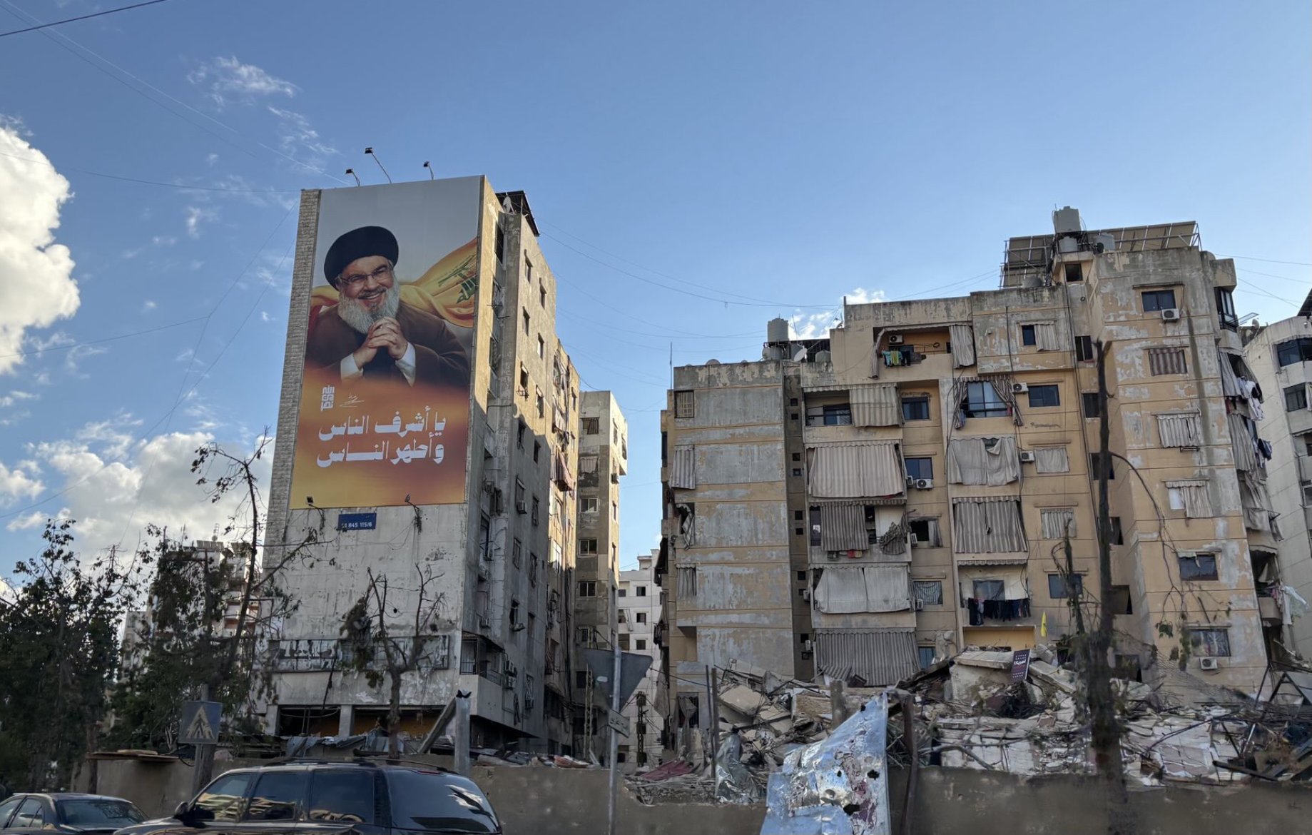 در این تصویر، یک ساختمان بزرگ با تصاویری از فردی که احتمالاً یک شخصیت عمومی است، دیده میشود. در کنار آن، ساختمانهای دیگری وجود دارند که به دلیل آسیبهای ناشی از زلزله یا جنگ، خرابه و آسیبدیده به نظر میرسند. وضعیت بناها نشاندهنده تخریب و ویرانی قابل توجهی است. آسمان در حال حاضر روشن است و ابرهای سفیدی در آن دیده میشود. سبزینگی در اطراف نیز وجود دارد که ممکن است درختان یا گیاهان باشد.