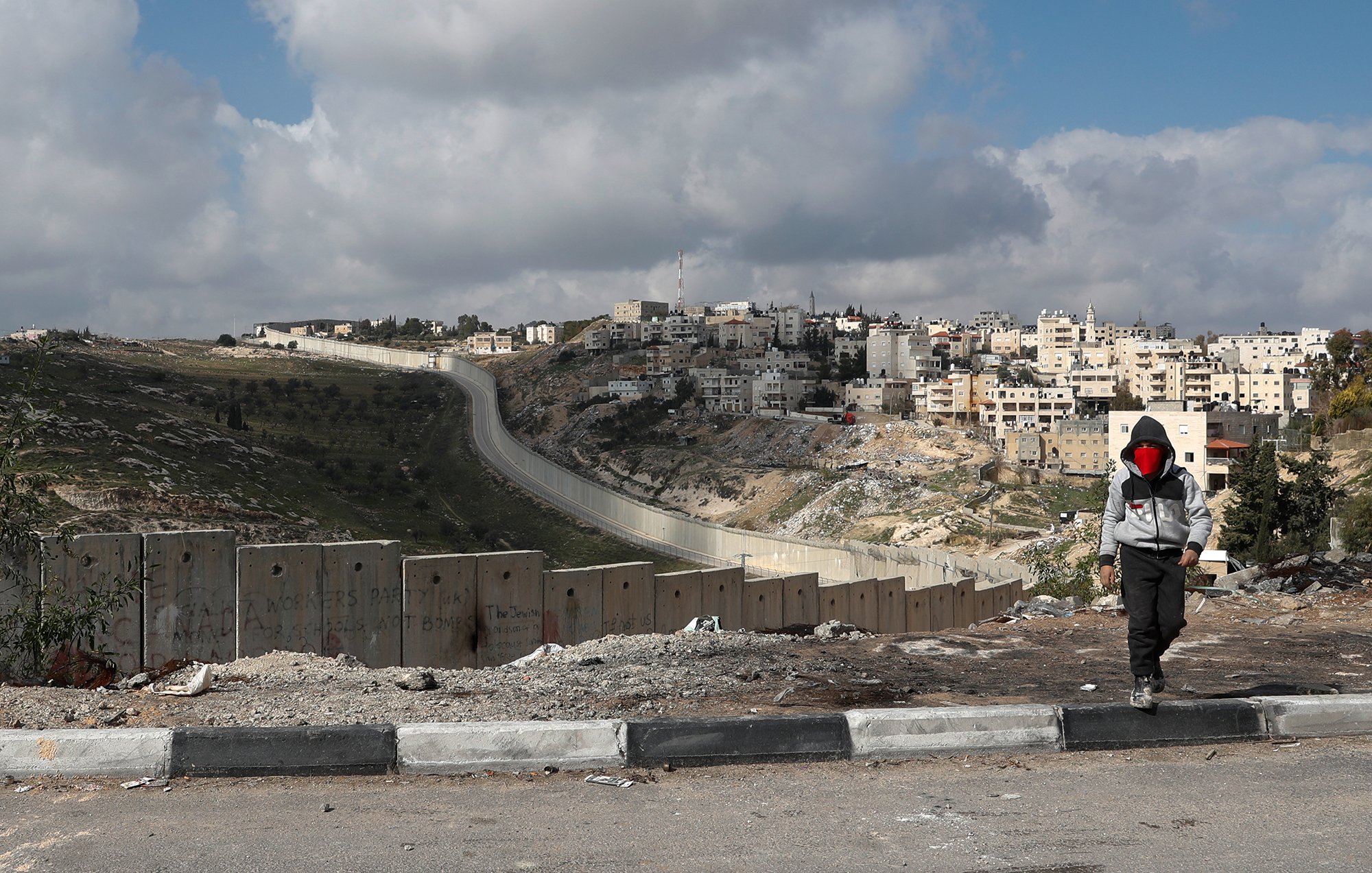 در این تصویر، یک پسر در حال پیادهروی در کنار دیواری بلند و سیمخاردار به نظر میرسد. این دیوار در زمینهی یک منطقه شهری قرار دارد که ساختمانهای مسکونی در آن دیده میشود. آسمان ابری است و ابرها در حال حرکت هستند. فضای تصویر ممکن است احساساتی از تنهایی و جدایی را منتقل کند. جادهای در جلوی تصویر وجود دارد و محیط بهنظر خشک و ناآرام میآید.