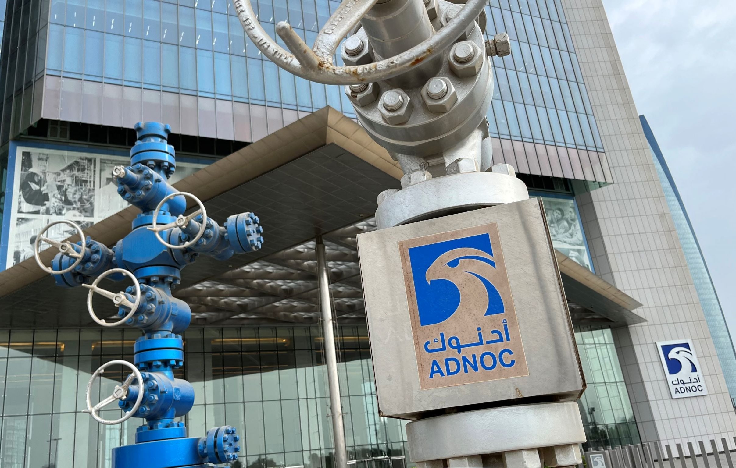 تصویر نشاندهنده نمای ساختمان شرکت نفت ابوظبی (ADNOC) است. در جلو، دو ساختار فلزی وجود دارد که شبیه به تجهیزات صنعتی و دکلهای نفتی هستند. یکی از این تجهیزات به رنگ آبی و دیگری نقرهای است. در سمت راست تصویر، لوگوی ADNOC با نوشته عربی و انگلیسی قابل مشاهده است. ساختمان دارای ظاهری مدرن و شیشهای است که عناصر طراحی معاصر را به نمایش میگذارد.