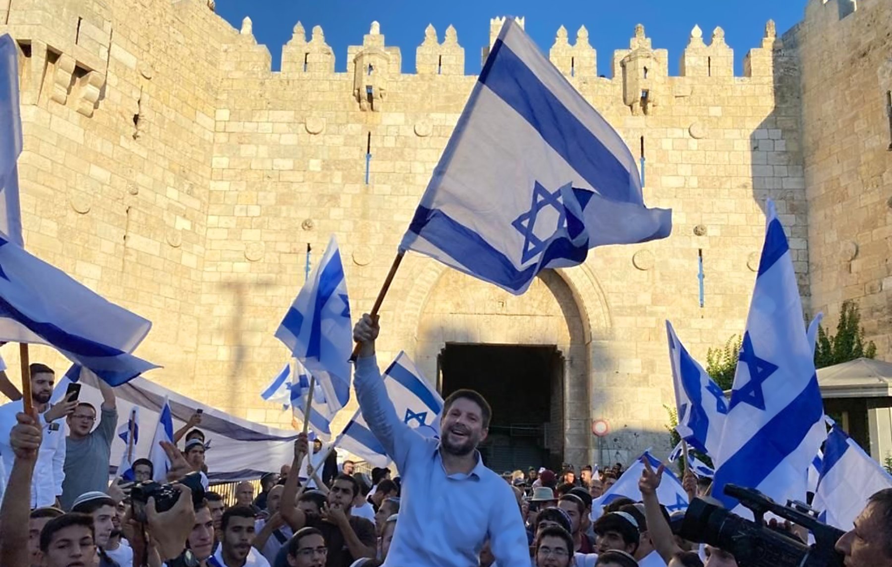 تصویر نشاندهنده جمعیتی است که با پرچمهای اسرائیل در حال جشن و شادی هستند. در میان این جمعیت، فردی در حال بلند کردن پرچم و ابراز شادی است. پسزمینه تصویر یک دروازه تاریخی و دیوار سنگی نمایان است که نشاندهنده یک مکان مهم است. افراد حاضر در تصویر لباسهای مختلفی به تن دارند و فضای کلی عکس نشاندهنده شور و نشاط است.