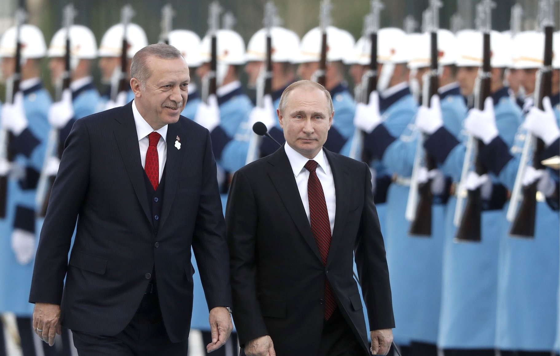 در این تصویر، دو فرد نزدیک به هم در حال راه رفتن هستند. یکی از آنها یک کت و شلوار تیره به همراه یک کراوات قرمز به تن دارد، و دیگری یک کت و شلوار تیره با کراوات مشکی پوشیده است. در پسزمینه، گروهی از سربازان در یونیفرمهای آبی روشن و سفید ایستادهاند که سلاحهای خود را در دست دارند. این تصویر شکلی از احترام و مراسم رسمی را نشان میدهد.