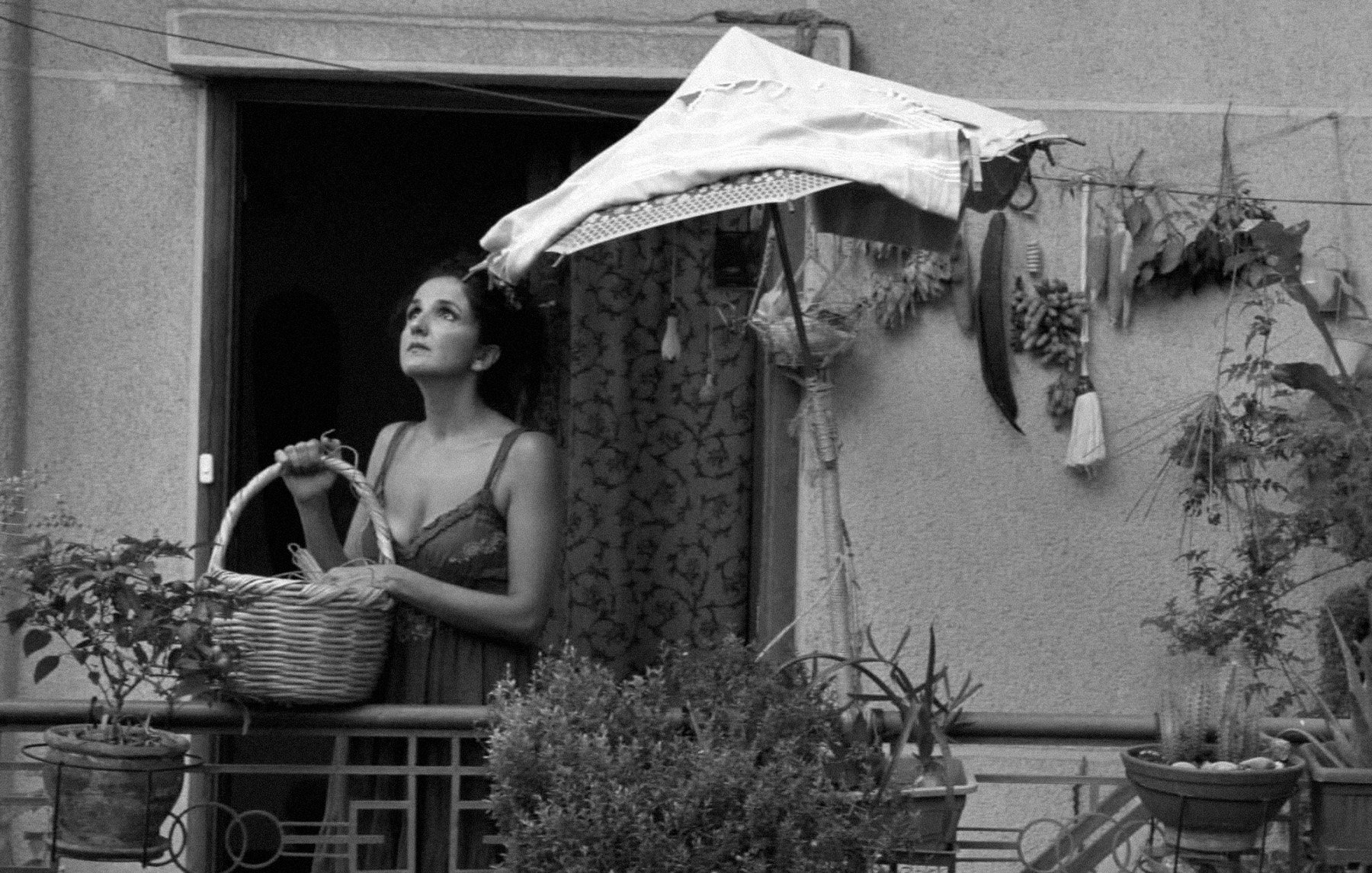 در تصویر، زنی در بالکن ایستاده و سبدی در دست دارد. او به نظر میرسد در حال نگاه کردن به بیرون است. بالکن پر از گیاهان و گلهاست و سایهبان کوچکی نیز بالای سرش وجود دارد. فضای تصویر بهصورت سیاه و سفید است که به آن حس قدیمی و عاشقانهای میدهد. دیوارهای اطراف ساده و بدون تزئینات هستند. این تصویر احساس تنهایی و تأمل را القا میکند.