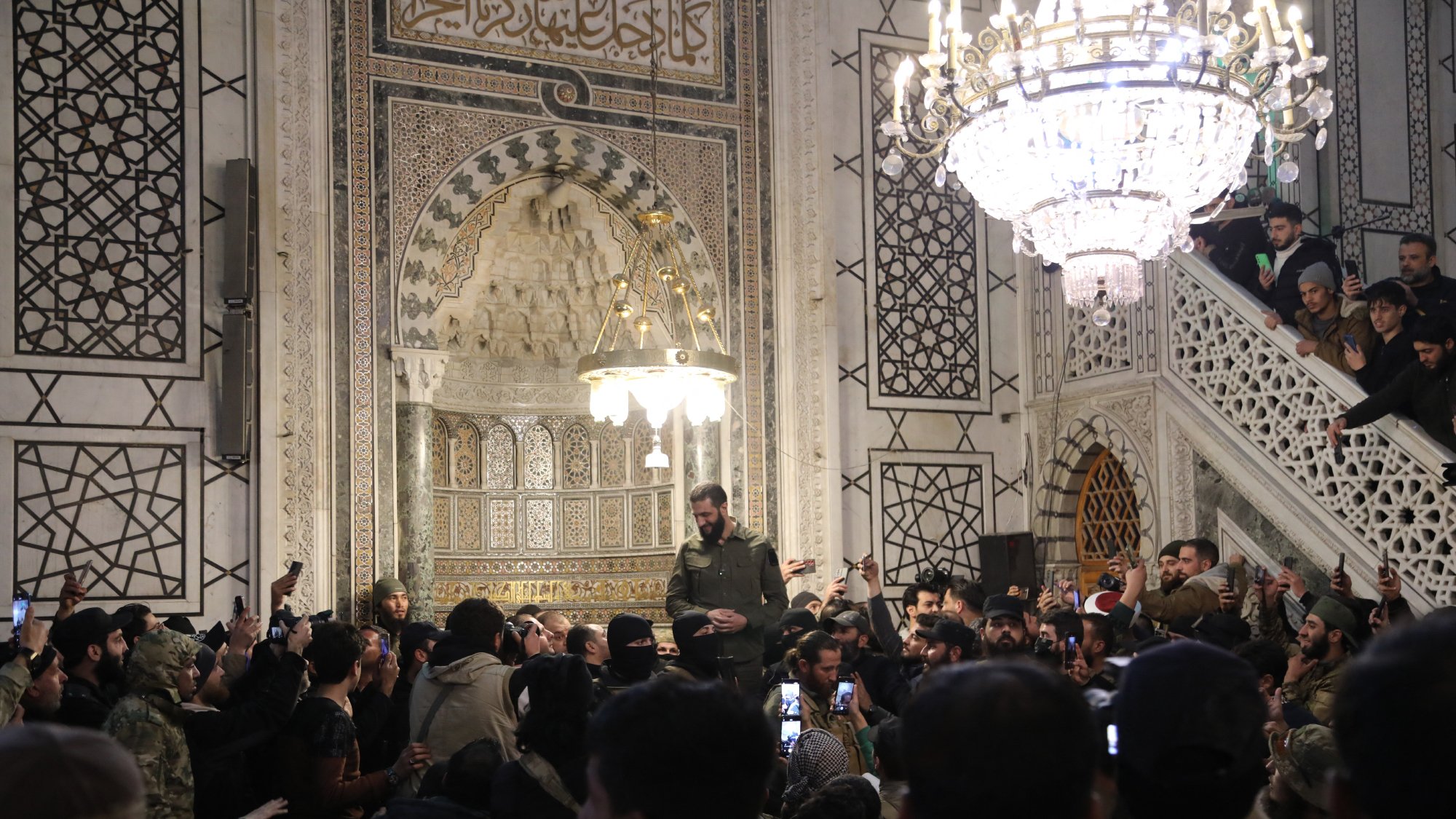 در تصویر، گروهی از افراد در یک مکان مذهبی یا تاریخی تجمع کردهاند. در وسط تصویر، فردی با لباس نظامی در حال سخنرانی یا بیان مطلبی به جمعیت است. نفرات در اطراف او با تلفنهای همراه خود مشغول فیلمبرداری یا عکاسی هستند. محیط اطراف آنها تزئینات زیبا و نورپردازی چشمگیری دارد، همچنین یک چراغ آویز بزرگ در بالای سر گروه دیده میشود که به فضا زیبایی میبخشد. دیوارها با نقوش و طراحیهای پیچیده تزئین شدهاند و جو عمومی تصویر نشاندهنده شور و شوق جمعیت است.