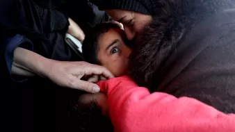 در این تصویر، یک زن در حال در آغوش گرفتن و بوسیدن یک کودک است. چهره کودک با نگاهی معصوم و کنجکاو به دوربین مینگرد. دست دیگری به آرامی به کودک نزدیک شده است. فضای تصویر احساسی و صمیمی به نظر میرسد، و ارتباط عاطفی بین مادر و فرزند در آن مشهود است. رنگهای لباسها و پوستهها، حس نزدیکی و حمایت را منتقل میکند.