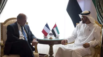 در این تصویر، دو نفر در یک محیط رسمی نشستهاند. یکی از آنها که لباس رسمی پوشیده است، به سمت چپ تصویر قرار دارد و به نظر میرسد مسئول یا مقام دولتی از فرانسه باشد. فرد دیگر که لباس سنتی عربی را بر تن دارد، به سمت راست تصویر نشسته و به نظر میرسد که مقام بلندپایهای از امارات است. در میان آنها، دو پرچم، یکی از فرانسه و دیگری از امارات متحده عربی، بر روی میزی کوچک قرار دارد. فضای اتاق، با پردههای زیبا و تزئینات سلطنتی، حاکی از یک ملاقات دیپلماتیک است.