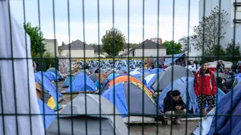 این تصویر نمایی از یک اردوگاه چادرها را نشان میدهد. چادرهای متعدد با رنگهای مختلف در یک فضای باز قرار دارند و برخی از افراد در اطراف آنها مشغول فعالیت هستند. در پسزمینه، یک دیوار با هنر خیابانی یا گرافیتی قابل مشاهده است. همچنین، یک نرده فلزی در جلوی تصویر وجود دارد که فضا را از محیط بیرونی جدا میکند. هوای آسمان میتواند کمی ابری به نظر برسد.