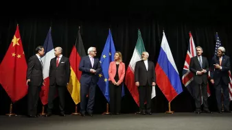 در این تصویر، گروهی از نمایندگان کشورهای مختلف در یک جلسه رسمی ایستادهاند. پرچمهای چین، آلمان، اتحادیه اروپا، ایران، روسیه، بریتانیا و ایالات متحده در پسزمینه حضور دارند. هر یک از افراد در لباسهای رسمی و با حالتهای متفاوتی در صف ایستادهاند، که نشاندهنده اهمیت و جدیت جلسه است. به نظر میرسد آنها در حال گفتوگو و تعامل با یکدیگر هستند.