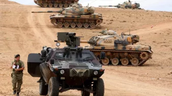 تصویر نشاندهنده یک صحنه نظامی است که در آن چندین تانک در حال حرکت یا منتظر هستند. در جلوی تصویر، یک خودرو نظامی سنگین قرار دارد که یک سرباز در حال بررسی یا نگهبانی در کنار آن ایستاده است. زمین کوهستانی و خشک به نظر میرسد و فضای کلی تصویر حس نظامی و جدی بودن را منتقل میکند. تانکها به سمت دورتر تصویر حرکت کرده و در حال آمادهباش به نظر میرسند.