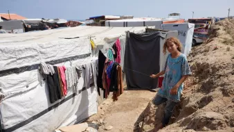 تصویر یک دختر بچه را در یک محیط غیررسمی نشان میدهد که احتمالاً یک اردوگاه پناهندگان یا محلی با شرایط زندگی سخت است. این دختر در کنار یک راه باریک ایستاده و لباسی ساده به تن دارد. در اطراف او، چادرها و سازههای موقت دیده میشود که لباسهایی به طناب آویزان شدهاند. زمین خاکی و نامنظم به نظر میرسد و فضا یک حس ناامنی را منتقل میکند. هوای آبی در پسزمینه نشان میدهد که این مکان نزدیک به دریا یا یک منبع آب است.