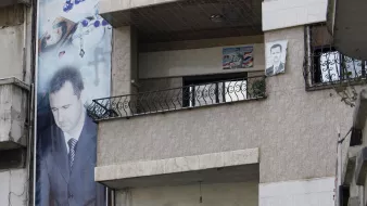 در این تصویر، نمایی از یک ساختمان چند طبقه دیده میشود که در آن پوسترها و تصاویر بزرگی از یک شخص (که احتمالاً یک شخصیت سیاسی است) نصب شده است. دو مرد در بالکن طبقه دوم ایستادهاند و به فضای بیرون نگاه میکنند. همچنین، در زیر ساختمان، بنرهایی با شعارها و تصاویری از دیگر افراد نصب شده است. محیط به نظر میرسد که در یک منطقه شهری باشد و برخی نشانههای ملی مانند پرچم در تصویر دیده میشود.
