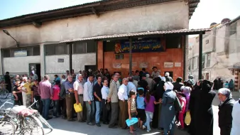 در تصویر، گروهی از مردم به صف ایستادهاند. ساختمان در پسزمینه دارای نمای سادهای است و تابلو بزرگی بالای درب ورودی نصب شده است. افراد در صف شامل مردان و زنانی هستند که برخی از آنها سبدها یا کیسههای خرید را در دست دارند. نور خورشید قوی به تصویر تابیده و فضا شلوغ به نظر میرسد. برخی از افراد به طرف دوربین نگاه میکنند و برخی دیگر مشغول صحبت هستند. دوچرخهها نیز در کنار خیابان به چشم میخورند.