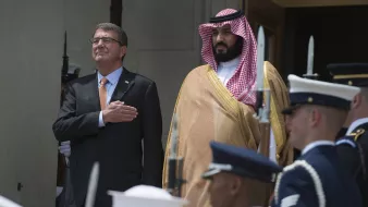 در این تصویر، دو مرد ایستادهاند. مرد سمت چپ با یک کت و شلوار تیره و پیراهن روشن دیده میشود و دستانش را بر روی سینهاش گذاشته است، که نشاندهنده احترام یا تقدیر است. مرد سمت راست در لباس رسمی عربی و با عمامهای که به طور خاص مرتب شده، stands. در پسزمینه، گروهی از سربازان در یونیفورمهای نظامی در حال ایستادن هستند. این صحنه احتمالاً به یک مراسم رسمی یا استقبال سیاسی مربوط میشود.