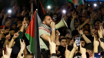تصویر نشاندهنده جمعیتی از افراد است که در یک تجمع اعتراضی شرکت کردهاند. در مرکز تصویر، مردی با میکروفون در دست در حال سخنرانی یا فریاد زدن است. این فرد با پوششی که شامل شال معروف فلسطینی است، به نظر میرسد که به موضوعی مهم صحبت میکند. در پسزمینه، جمعیت زیادی با پرچمهای فلسطین و دستهایی که به نشانه اعتراض یا حمایت بالا رفتهاند، دیده میشود. این تصویر وضعیت هیجانانگیز و پرشور این تجمع را منتقل میکند.