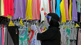در این تصویر، یک زن با حجاب در حال خریداری لباس از یک فروشگاه است. او دستکش به دست دارد و صورتش را با یک محافظ شفاف پوشانده است. لباسهای متنوعی با رنگهای مختلف مانند زرد، بنفش و آبی در پشت او آویزان شدهاند. زن در حال بررسی لباسها و انتخاب آنها به نظر میآید.