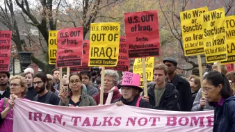در این تصویر، گروهی از معترضان در حال برگزاری یک تظاهرات هستند. آنها پلاکاردهایی با نوشتههای مختلف در دست دارند که بر ضد دخالتهای نظامی ایالات متحده در عراق، سوریه و افغانستان و همچنین بر ضد تحریمها و جنگ تأکید میکند. همچنین، در میان جمعیت، یک بنر بزرگ با نوشته «ایران، گفتگو نه بمب» مشاهده میشود. رنگ بیشتر پلاکاردها زرد است و جمعیت نشان از تنوع دارد. فضای تظاهرات جدی و متعهدانه به نظر میرسد.