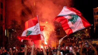 در تصویر، جمعیتی از مردم در یک تظاهرات شبانه دیده میشوند. افراد با پرچمهای لبنان در دست، در حال شادی و ابراز احساسات هستند. یک شعله آتش بزرگ در پسزمینه به روشنایی فضای شب افزوده و به جو فعالیت و هیجان دامن میزند. تعداد زیادی از افراد گوشیهای هوشمند خود را برای ثبت لحظات بالا بردهاند. این صحنه نمایانگر شور و شوق اجتماعی و همبستگی مردم است.