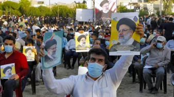 در تصویر، جمعیتی از افراد در یک فضای باز دیده میشود که روی صندلی نشستهاند و تعدادی از آنها در حال نگهداشتن پوسترهایی از یک شخصیت سیاسی هستند. مردم ماسک بهصورت دارند و در پسزمینه، تعدادی پرچم و تابلوهای نمایشی نیز وجود دارد. این جمعیت به نظر میرسد که در یک رویداد یا حمایت سیاسی شرکت کردهاند.