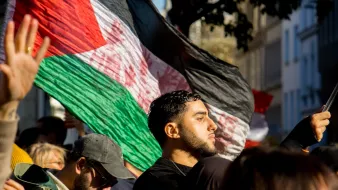 در این تصویر، گروهی از افراد در حال برگزاری یک تظاهر یا تجمع هستند. یک مرد جوان در جلو تصویر دیده میشود که به پرچم کشور خود (که احتمالاً پرچم فلسطین است) افتخار میکند. او با جدیت به جلو نگاه میکند. در پسزمینه، تعدادی از افراد دیگر نیز حضور دارند که برخی از آنها کلاه به سر دارند و به نظر میرسد که در حال حمایت از یک هدف مشترک هستند. نور آفتاب به تصویر زیبایی لطافتی میبخشد و فضای تجمع را گرم و زنده نشان میدهد.