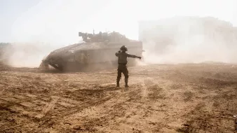 تصویر نشاندهندهی یک صحنهی نظامی است که در آن یک تانک در محیطی گرد و خاکی قرار دارد. نظامیای که در جلو دیده میشود، به سمت دیگری اشاره میکند و به نظر میرسد در حال هدایت یا مدیریت تحرکات است. نور شدید و شرایط گرد و خاکی نمایانگر یک فضای پرتنش و عملیاتی است. در پسزمینه، میتوان ساختمانهایی را مشاهده کرد که به سختی مشخص هستند.