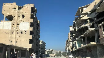 تصویر یک خیابان خالی را نشان میدهد که در دو طرف آن ساختمانهای مخروبه و آسیبدیده قرار دارند. دیوارهای این ساختمانها پر از سوراخها و آسیبهای ناشی از جنگ یا تخریب هستند. در انتهای خیابان، چند ساختمان دیگر نیز مشاهده میشود. آسمان آبی و صاف است و افراد کمی در خیابان در حال تردد هستند. دو نفر در سمت چپ تصویر به نظر میرسد در حال صحبت کردن هستند و یک کودک نیز با آنهاست. فضای کلی تصویر تنهایی و تخریب را القا میکند.