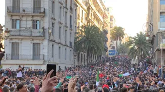 این تصویر نمایی از یک اجتماعات بزرگ در خیابانی شلوغ را نشان میدهد. مردم زیادی در حال تظاهرات یا جمع شدن هستند و در میان آنها تعدادی پرچم به چشم میخورد. درختان بلند نخل در دو طرف خیابان دیده میشوند و ساختمانهای مرتفع با بالکنها نیز در پسزمینه قرار دارند. مردم با entusiasmo در حال برنداشتن تلفنهای همراه خود و ثبت این لحظه هستند. جو عمومی تصویری از شور و شوق و همبستگی را القا میکند.