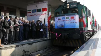 در این تصویر، یک قطار در ایستگاهی با مقامات و افراد حضور دارد. در پسزمینه، پرچمهای کشورهای مختلف به چشم میخورد. چندین نفر در سمت چپ تصویر ایستاده و به سمت قطار توجه دارند، برخی از آنها در حال applauding هستند. تابلوهایی با متنهایی درباره افتتاح و همکاریهای بینالمللی نیز در تصویر دیده میشود. این صحنه نشاندهنده یک رویداد ویژه یا افتتاحیه مرتبط با حمل و نقل ریلی است.