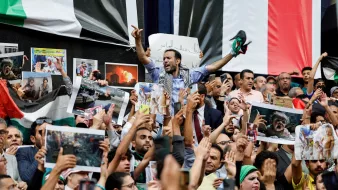 این تصویر یک تجمع بزرگ را نشان میدهد که مردم در آن با شور و هیجان حضور دارند. تعدادی از افراد در حال نشان دادن تصاویر و پلاکاردهایی هستند. برخی از معترضان در حال فریاد زدن یا ابراز احساسات خود هستند و مشخص است که موضوعی برای آنها اهمیت دارد. در پسزمینه، پرچمها و تصاویری از وقایع یا شخصیتهای مهم ممکن است مشاهده شود. جو کلی تصویر پر از انرژی و احساسات قوی است.