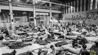 این تصویر یک فضای داخلی را نشان میدهد که پر از تختهای بیمارستانی است. افراد مختلفی در حالتهای مختلف دراز کشیدهاند، برخی در حال استراحت و برخی دیگر در حال حرکت یا صحبت با یکدیگر هستند. محیط به نظر شلوغ و پر از فعالیت میرسد، و پوشش بیشتر افراد ساده و شامل چادر یا پتو است. دیوارها به نوعی دکور شدهاند، و فضای کلی شاید به یک مرکز درمانی یا پناهگاه اشاره دارد.