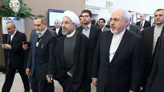 این تصویر گروهی از افراد را نشان میدهد که در حال حرکت به سمت یک مکان هستند. یکی از آنها به وضوح لباس روحانیت پوشیده و دیگران نیز به طور رسمی لباس پوشیدهاند. به نظر میرسد این افراد در یک رویداد یا کنفرانس رسمی شرکت کردهاند. محیط تصویر ممکن است یک مکان اداری یا کنفرانس باشد که نشاندهنده تعاملات سیاسی یا اقتصادی است.