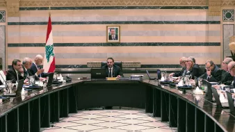 تصویر یک جلسه رسمی دولتی در لبنان را نشان میدهد. تعدادی از مقامات و اعضای دولت در اطراف یک میز بزرگ نشستهاند. در مرکز جلسه، یک مرد با لباسی رسمی و آرامش بر روی صندلی ریاست نشسته است و به دیگران نگاه میکند. در پسزمینه، پرچم لبنان و تصویری از یک شخصیت مهم بر دیوار نصب شده است. فضای اتاق با خطوط عمودی و افقی تزئینی و رنگهای ملایم طراحی شده است. افراد حاضر در جلسه به موضوعات مختلفی مشغول بحث و تبادل نظر هستند.