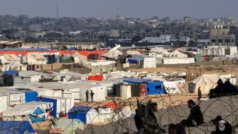 این تصویر منطقهای را نشان میدهد که پر از چادرهای سفید و آبی است. در پسزمینه، ساختمانهای شهری دیده میشود. جمعی از افراد در نزدیکی چادرها نشستهاند و به نظر میرسد که در یک وضعیت دشوار زندگی میکنند. همچنین، نوارهایی از سیمخاردار در عکس به چشم میخورد که نشاندهنده محدودیتهای امنیتی است. فضای کلی تصویر حاکی از چالشهای انسانی و وضعیت اضطراری است.