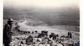 در این تصویر، مردی با کلاه و پوشاک سنتی در حال ایستادن در کنار یک شیب سنگی دیده میشود. در پسزمینه، گروهی از انسانها و حیوانات (احتمالاً خرها یا اسبها) در حال حرکت در یک مسیر سنگی به سمت پایین هستند. مناظر اطراف خشک و کوهستانی به نظر میرسد و تصویر حالتی قدیمی و سیاه و سفید دارد که به آن احساس تاریخ را القا میکند. فضای تصویر و نوع پوشش افراد نشاندهنده یک فعالیت یا سفر است.