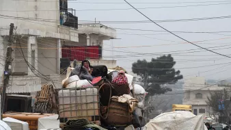 در این تصویر، گروهی از افراد در حال سفر بر روی یک کامیون بار هستند. آنها به طور واضحی در میان بار زیادی از محصولات و وسایل قرار دارند. برخی از افراد پوشیده از لباسهای ضخیم و شالهای رنگی به نظر میرسند که به دلیل سرما به خود پیچیدهاند. محیط اطراف شلوغ و شهرنشین به نظر میرسد، با ساختمانها و خطوط برق مشخص. وضعیت کلی تصویر احساس چالش و سختی را القا میکند.