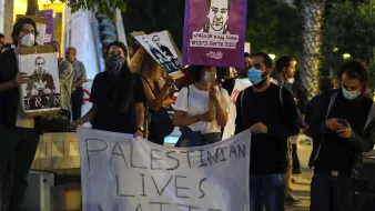 در این تصویر، گروهی از مردم در حال برگزاری یک تجمع اعتراضی هستند. آنها تابلوهایی با تصاویر و شعارهایی در دست دارند. یکی از بنرهای بزرگ به زبان انگلیسی میگوید "زندگیهای فلسطینی مهم است". شرکتکنندگان به وضوح از ماسک استفاده میکنند، و فضای عمومی تجمع نشاندهنده یک اعتراض اجتماعی است. در پسزمینه، درختان و یک محیط شهری قابل مشاهده است.