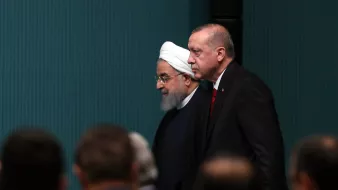 در تصویر، دو شخصیت سیاسی در حال حرکت به سمت جلو دیده میشوند. یکی از آنها لباس سنتی با عمامه سفید به سر دارد و دیگری کت و شلوار تاریک و کراواتی بر تن دارد. پسزمینه تصویر و دیگر افراد حاضر در جلسه به خوبی واضح نیستند و تمرکز بر روی این دو شخصیت است. محیط به نظر میرسد که رسمی باشد.