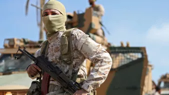در این تصویر، یک سرباز مسلح با پوشش صورت، در حال ایستادن در پیشزمینه قرار دارد. او یونیفورم نظامی و کلاهی به سر دارد و یک اسلحه در دستش است. در پسزمینه، یک خودروی نظامی دیده میشود و تعدادی از سربازان دیگر در حال فعالیت هستند. آسمان آبی و آب و هوای روشن، صحنه را روشن میکند. این تصویر حس قدرت و دقت نظامی را به تصویر میکشد.