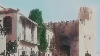 در این تصویر تاریخی، یک گروه از نظامیان در حال عبور از خیابانی در شهری با معماری قدیمی هستند. در دو طرف خیابان، جمعیتی زیادی ایستادهاند و به تماشای اتفاقات میپردازند. در پسزمینه، ساختمانهایی با طراحی خاص و دیواری بزرگ دیده میشوند. برخی از افراد در بالای دیوار ایستادهاند و نظارهگر هستند. فضای تصویر حسی از یک رویداد رسمی یا مراسم ویژه را القا میکند و رنگآمیزی تصویر به آن جذابیت بیشتری بخشیده است.