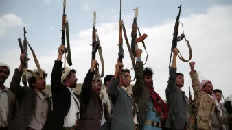 در تصویر، گروهی از مردان با لباسهای سنتی و محلی در حال ایستادن هستند و تفنگهایی را در دستان خود بالا بردهاند. آنها در حال نشان دادن اتحاد و یا ابراز قدرت خود هستند. پسزمینه آسمانی با ابرهای کمرنگ دیده میشود و حالت کلی تصویر بیانگر یک وضعیت احساسی و ممکن است در حال برگزاری یک تجمع یا مراسم خاص باشد.