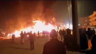 در این تصویر، ظاهراً افرادی در یک صحنه شبانه به سوی یک آتش بزرگ در حال تماشای آن هستند. شعلههای آتش به شدت در حال سوزاندن یک ساختمان یا ساختار دیگر هستند و دود سیاهی به سمت آسمان بلند میشود. مردم در گروههای مختلف در پسزمینه ایستادهاند و به سمت محل آتش نگاه میکنند. احتمالاً این صحنه به یک حادثه یا اعتراض مربوط میشود. روشنایی آتش فضای اطراف را درخشان کرده است.