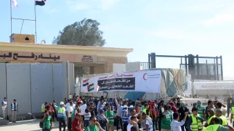 در تصویر، تعدادی از افراد در حال برگزاری یک تجمع یا اعتراض هستند. مردم در جلو ورودی یک ساختمان بزرگ ایستادهاند و برخی از آنها جلیقههای شرایطی به تن دارند. در پسزمینه، بنری بزرگ با متنی به زبان عربی و تصاویری از علمها به چشم میخورد. همچنین در بالای این ساختمان، علمهای کشورها در اهتزاز هستند. فضای تجمع به نظر شلوغ و پر انرژی میرسد.