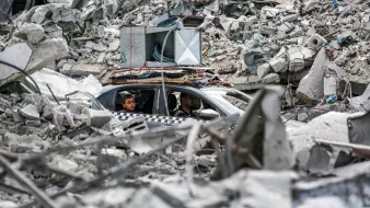 در تصویر، دو مرد درون یک خودرو در میان خرابهها نشستهاند. اطراف آنها پر از آوار و بقایای سازههای مخروبه است. برخی از أجزاء ساختمانی به وضوح دیده میشوند و وضعیتی به شدت آسیبدیده را نشان میدهند. در بالای خودرو، تجهیزات صوتی قرار گرفته است. فضای کلی تصویر حسی از ویرانی و بحران را منتقل میکند.