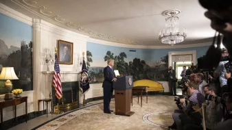 در این تصویر، دونالد ترامپ در حال سخنرانی است و در پسزمینه، دیواری با نقاشیهای منظره و یک پرچم ایالات متحده و یک پرچم دیگر دیده میشود. او در کنار یک تریبون ایستاده و دوربینهای خبرنگاران و عکاسان در جلو او قرار دارند. دکوراسیون اتاق زیبا و مجلل است، با یک فرش بزرگ و مجسمههایی در اطراف. نور طبیعی از پنجرهها به داخل اتاق میتابد.