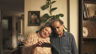 در این تصویر، یک زن و یک مرد در یک محیط داخلی ایستادهاند. زن لباسی با دکمههای بزرگ و کلاهی زیبا به تن دارد و در حال نشان دادن چیزی به مرد با گوشی همراه است. مرد در کنار او ایستاده و به صفحه گوشی نگاه میکند. هر دو لبخند بر لب دارند و به نظر میرسد که لحظهای خوشحالکننده را تجربه میکنند. پسزمینه تصویر نشاندهنده یک خانه گرم و دلنشین است با گیاهانی در کنار دیوار.