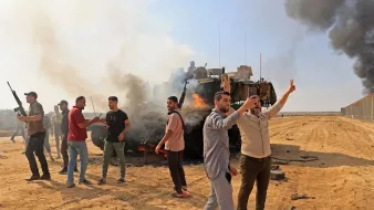 در تصویر، تعدادی از افراد در یک منطقه بیابانی مشاهده میشوند که به سمت دوربین اشاره میکنند و برخی از آنها سلاح بهدست دارند. در پسزمینه، دود سیاه و غلیظی از یک وسیله نقلیه نظامی در حال سوختن دیده میشود. به نظر میرسد که این صحنه به یک وقایع پرتنش یا بحران های اجتماعی مربوط میشود. افراد در حال ابراز هیجان و یا اعتراض هستند و فضای کلی تصویر، حالتی داغ و ناآرام دارد.