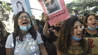 در این تصویر، گروهی از زنان و مردان در حال برگزاری یک تظاهرات هستند. آنها پلاکاردهایی در دست دارند که تصاویری از افراد مختلف بر روی آنها چاپ شده است. برخی از شرکتکنندگان به شدت فریاد میزنند و احساسات خود را ابراز میکنند. اکثر افراد ماسک به چهره دارند، که به خاطر وضعیت بهداشتی ممکن است باشد. فضای اطراف شلوغ به نظر میرسد و به وضوح نشاندهنده یک حرکت اجتماعی است.