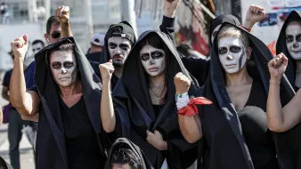 در این تصویر، گروهی از افراد زن و مرد را مشاهده میکنیم که لباسهای سیاهی به تن دارند و صورتهای خود را با رنگ سفید نقاشی کردهاند. آنها در حال نمایش اعتراض خود هستند و برخی از آنها با دستان بلند شده، نشانهای از قدرت و اتحاد را نشان میدهند. پسزمینه تصویر به وضوح حاکی از یک تجمع یا تظاهرات است. حالت چهرهها جدی و معنادار به نظر میرسد، که بیانگر عزم و اراده آنها برای بیان دیدگاهها و خواستههایشان است.