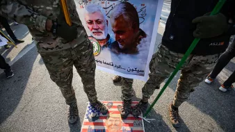 در این تصویر، تعدادی از افراد با لباسهای نظامی و camo (رنگهای استتاری) در حال ایستادن هستند. آنها در دست خود عصا یا چوبهایی دارند. زیر پایشان تصویر یک پرچم آمریکاست که بر روی آن چهرهای در پایین مشاهده میشود. در پسزمینه، یک بنر بزرگ وجود دارد که شامل تصاویری از دو نفر است و متنی در مورد آنها نوشته شده است. فضایی حاکی از تظاهرات یا یک رویداد اجتماعی در حال شکلگیری است.