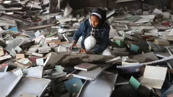 تصویر یک اتاق شلوغ و پر از کتابهای پراکنده است. یک دختر جوان در حال نشسته بر روی زمین، در حال بررسی یا جستجوی چیزی در میان کتابها و وسایل دیگر است. او لباسهای سادهای به تن دارد و فضای اطرافش بینظم و به هم ریخته به نظر میرسد.