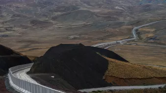 این تصویر نمایی از یک جاده در میان مناظر طبیعی خشک و کوهستانی را نشان میدهد. جاده به صورت منحنی و پیچدرپیچ از روی زمینهای ناهموار عبور میکند و در سمت راست تصویر، دیواری سفیدرنگ وجود دارد که به طور راستا در حال پیشروی است. در پسزمینه، تپهها و دامنههای کوهستانی با رنگهای ملایم و خاصی دیده میشوند که نمایی طبیعی و بکر را به تصویر میبخشند. فضای کلی تصویر به نوعی حس دورافتادگی و خلوتی را منتقل میکند.